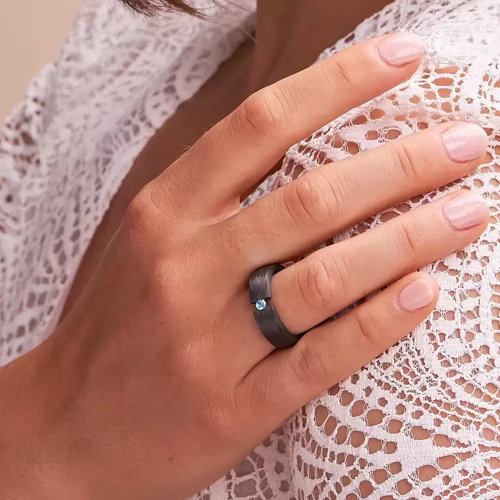 Carbon Ehering mit eingespanntem blauen Diamanten auf Hand an Schulter im Zoom